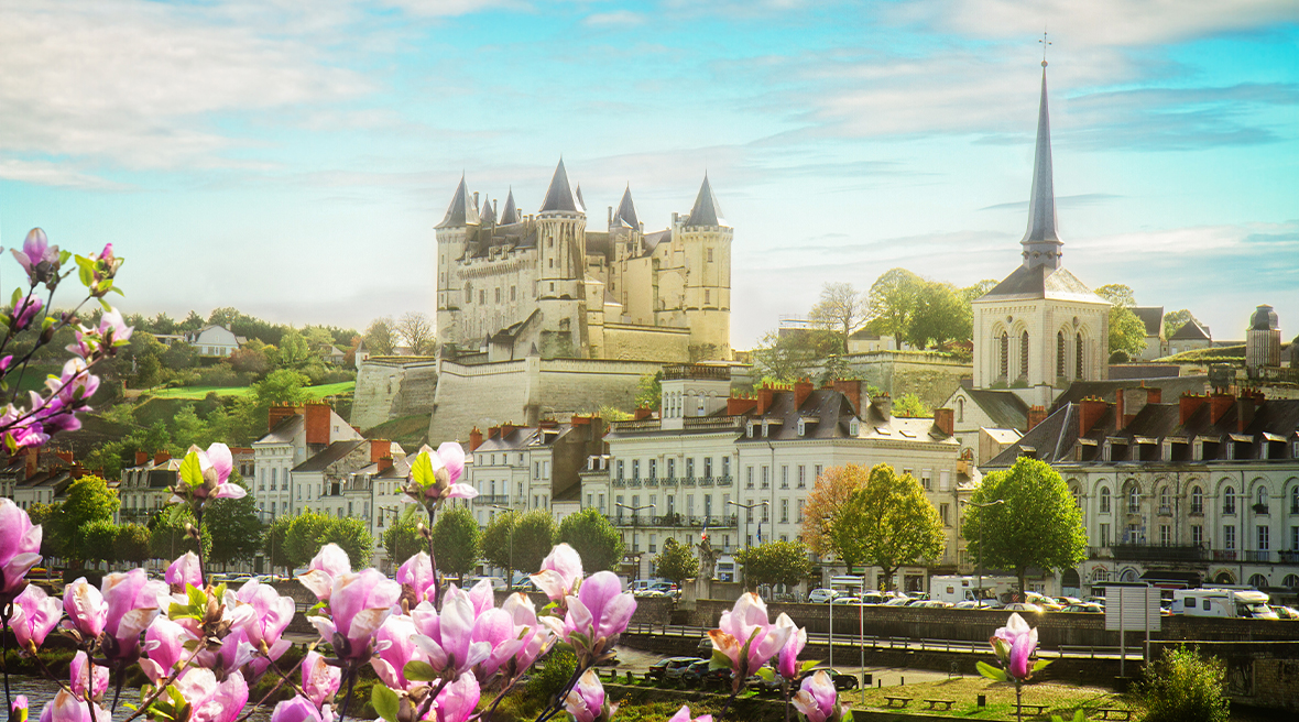 Romantic castle sits high above a row of buildings on a town’s river bank, with a church spire also prominent. Pink flowers in foreground